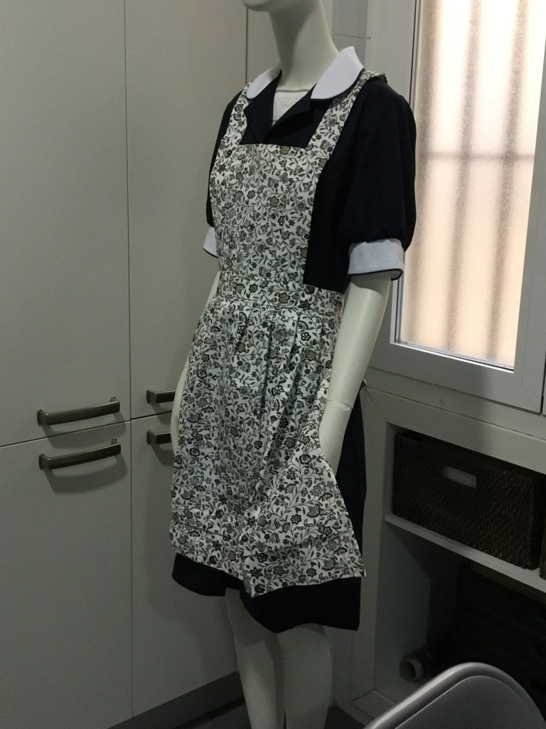 Uniforme para empleada del hogar formado por bata en color negro con detalles en blanco, y delantal floreado, de HOME&HOPE MADRID